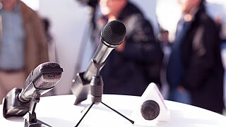 Close-up of two microphones, in the background is a blurred image of a man in suit and tie